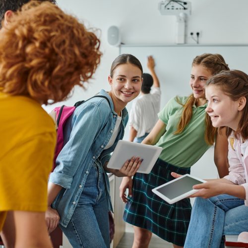 Smiling teen schoolgirls holding digital tablets near blurred friends in classroom in school